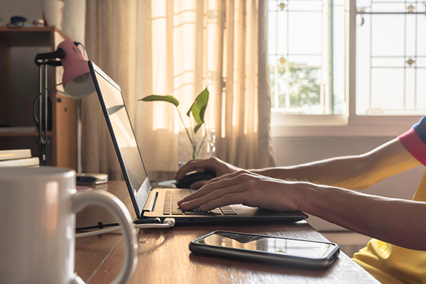 A person works from home at their computer desk, with a plant and a cup of coffee.