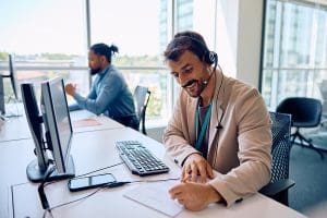 Happy call center agent taking notes while working on computer in office.
