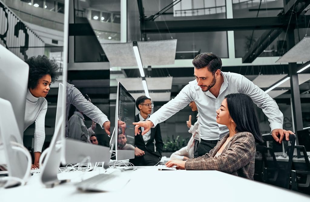 A team of IT experts in an open-plan office discuss information displayed on their computer screens.