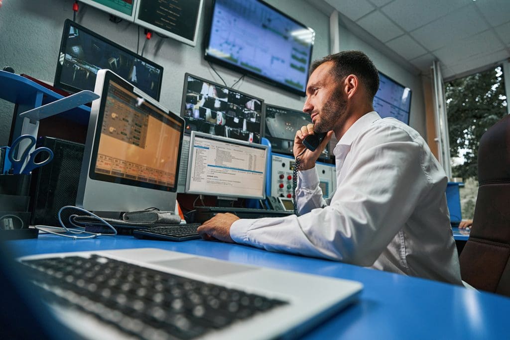 A man in an office with many computer screens holds a phone to his ear as he stares intently at a screen.